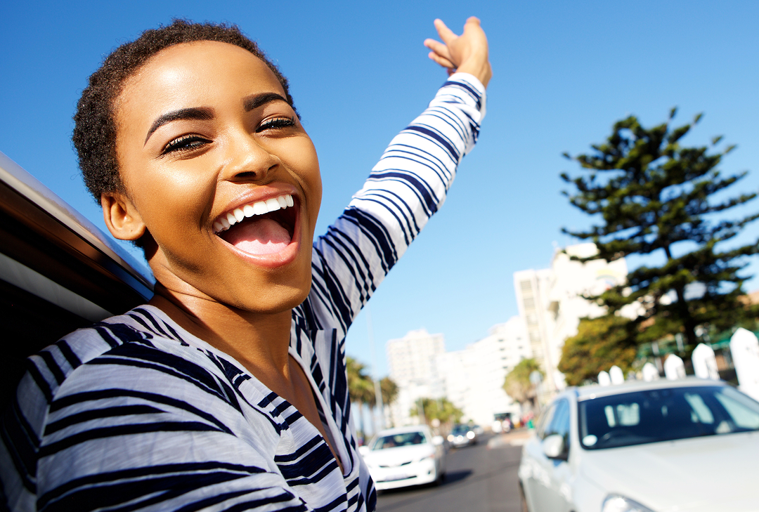 A smiling woman wearing a black and white shirt with her arm in the air. Blue sky and a tree appear in the background indicating that she is outside.